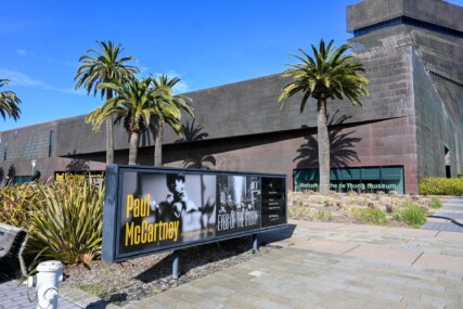 A view of the de Young Museum on a sunny day. The museum is in the background with a promo for a Paul McCartney exhibition in the foreground.