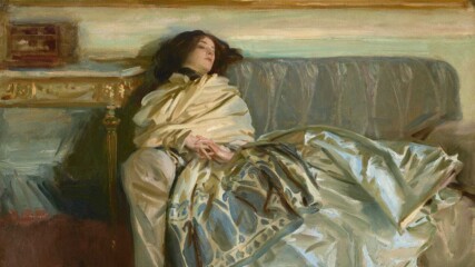 A woman with pale skin and dark brown hair is swathed in silky fabric as she reclines along a gray couch in this horizontal painting. Her head rests along the backrest of the sofa to our left, so her body extends to our right. Her torso is wrapped in a voluminous, ivory-white scarf or wrap. The scarf wraps tightly around her neck, and the bottom edge, near her knees, has a wide, indigo-blue pattern of ovals and vegetal forms. Her shimmering silver gray skirt is painted loosely with baby and sky blue, olive and sage green, and white strokes, and it drapes over the cushions and down the front of the couch. Her hands are clasped so her interwoven fingers rest at her navel. She looks down and off to our right. A table to our left is edged with gold. The wall behind her, above the couch, is painted with long streaks of eggshell white and pale turquoise. The gold frame of a dark painting hanging over the couch spans the width of the composition. Near the upper right corner of the canvas, the artist included a signature and as if he had signed the painting within this painting with loose, dark letters: “John S. Sargent 1911."